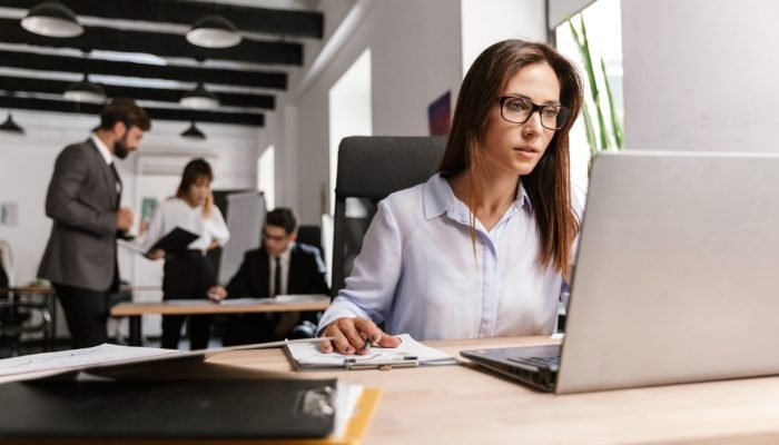 Photo of serious businesswoman wearing eyeglasses working with documents and laptop while sitting at desk in open-plan office